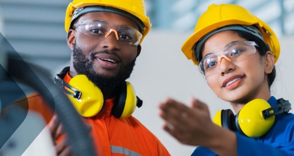 Engenheiros, um homem e uma mulher, sorrindo e usando equipamentos de proteção individual (EPIs), como capacetes e óculos de segurança.