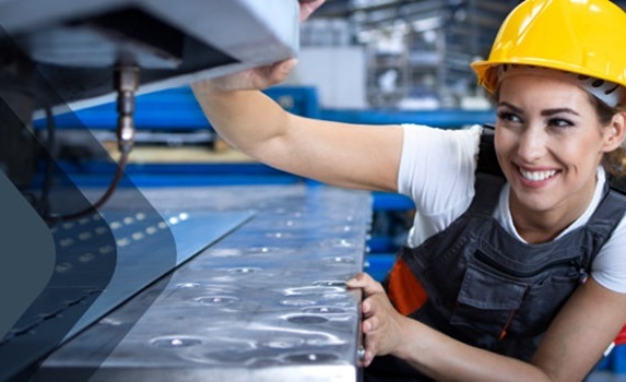 Mulher sorrindo, de capacete de segurança amarelo e uniforme, operando uma máquina em um ambiente industrial.