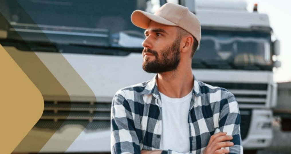 Motorista de caminhão com boné e camisa xadrez, de braços cruzados, em pé em frente a um caminhão.